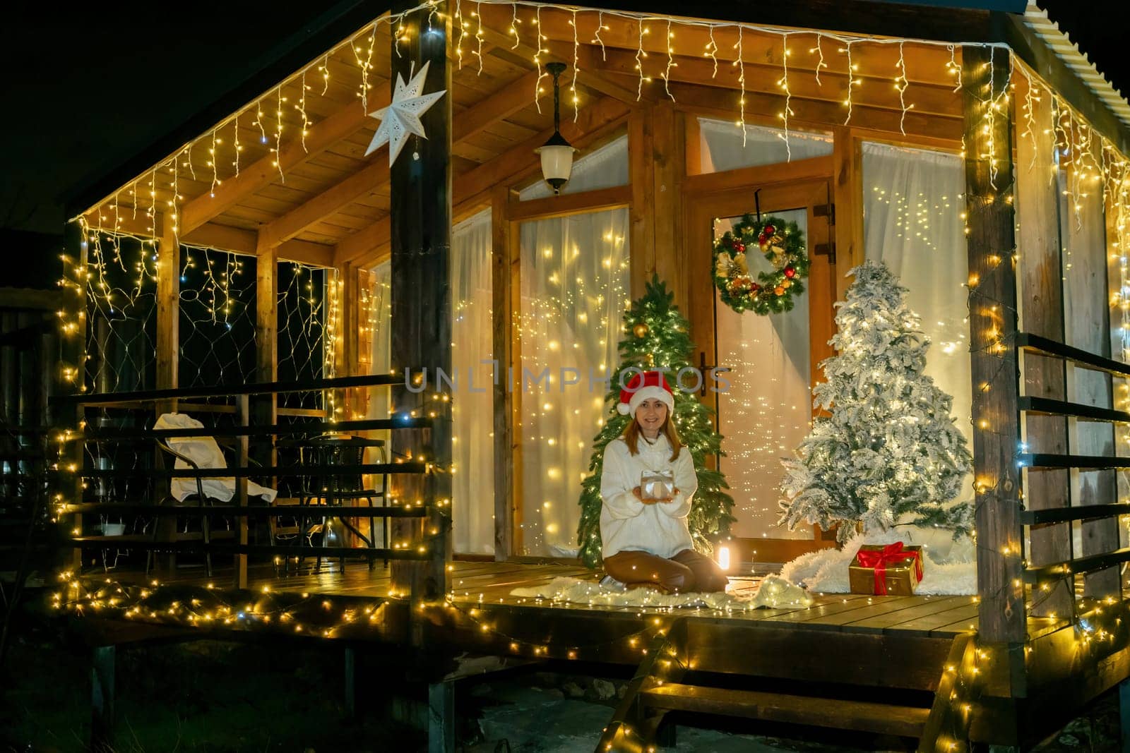 A decorated wooden cabin with a Christmas tree and string lights. A person wearing a Santa hat is seated on a bench, with a gift next to the tree.