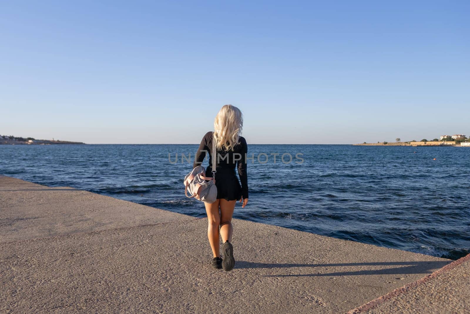 A woman with long blonde hair walks on a concrete pier overlooking the blue ocean. She wears a black dress and carries a large bag. The sky is bright blue and the sea is calm.