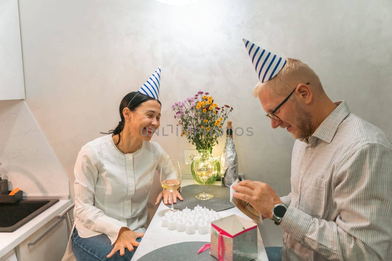 Here's a caption under 150 characters describing the image: Happy couple celebrates birthday in kitchen. They wear party hats and playfully interact, possibly opening gifts. A flower vase sits nearby.