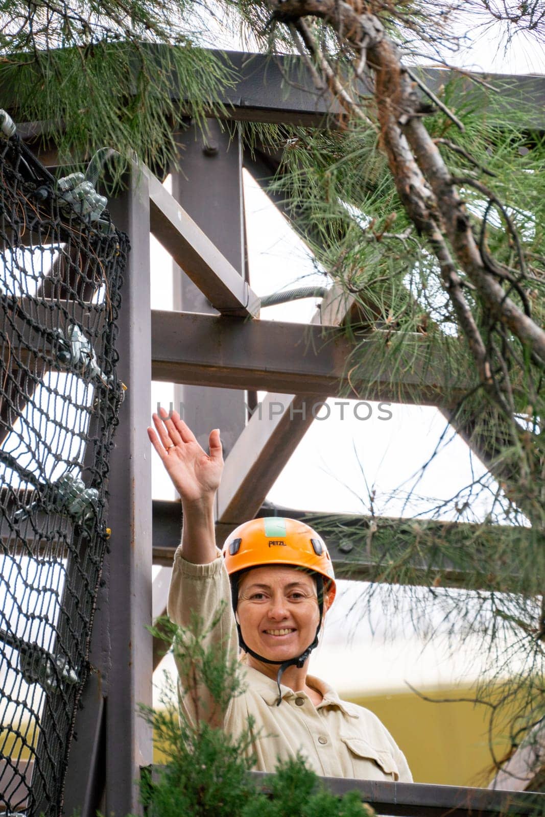 A woman wearing a helmet and khaki shirt smiles and waves from a wooden platform in a treetop obstacle course.