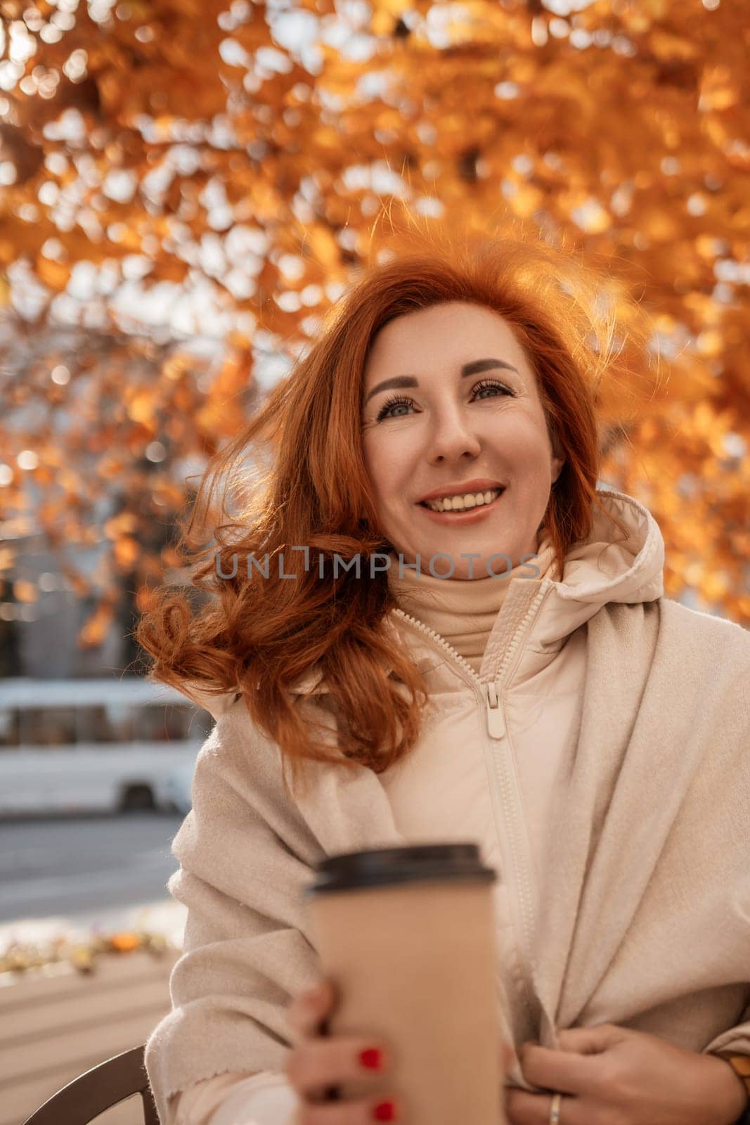 Smiling redhead woman, wrapped in a beige shawl, holds a coffee cup outdoors under a vibrant autumn tree. Golden leaves frame her.