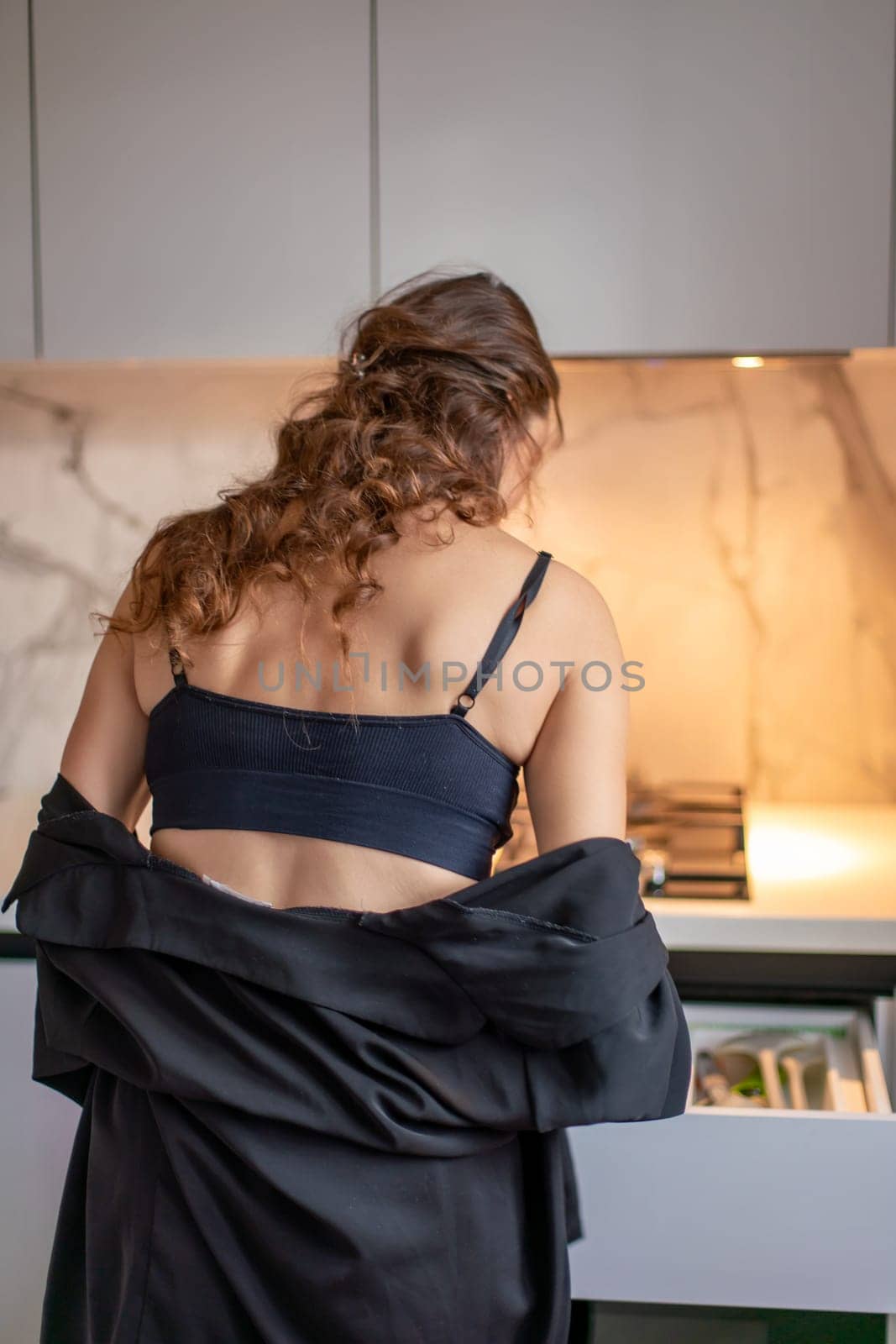 Woman with brown hair, wearing a black bra and robe, stands in a modern kitchen, back to the camera, partially opening a drawer.