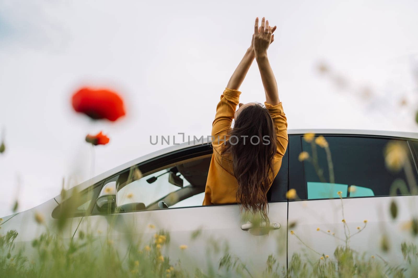 A woman is sitting in a car with her hands on her head, looking out the window. The car is parked in a field of flowers, with a red flower in the foreground. Concept of relaxation and freedom