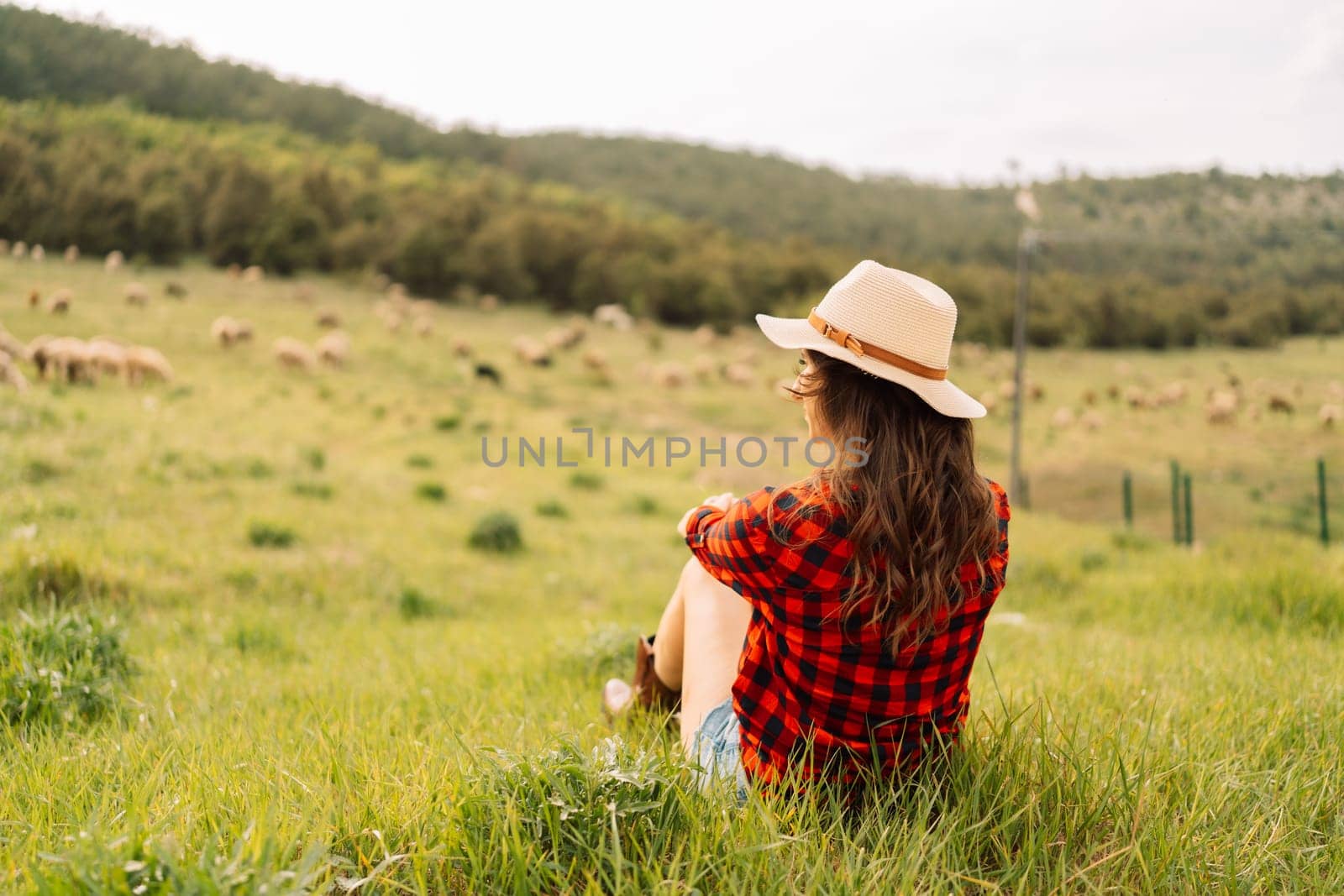 A woman wearing a straw hat and a red plaid shirt sits in a field of grass. The scene is peaceful and serene, with the woman looking out over the grassy field