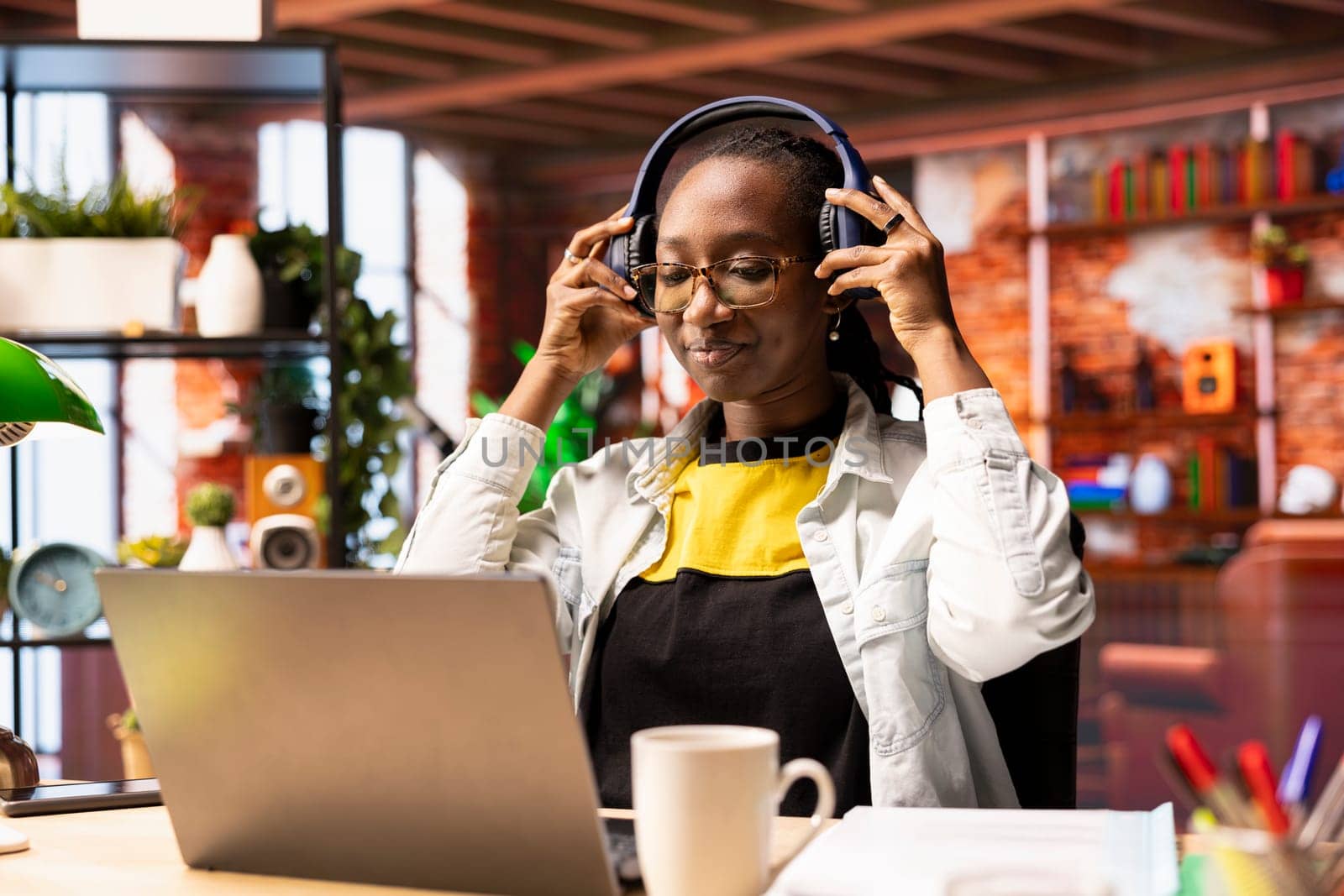 Cheerful woman in modern apartment room listening music during leisure time, feeling pleased. Joyous african american person at home hearing exciting songs through headphones