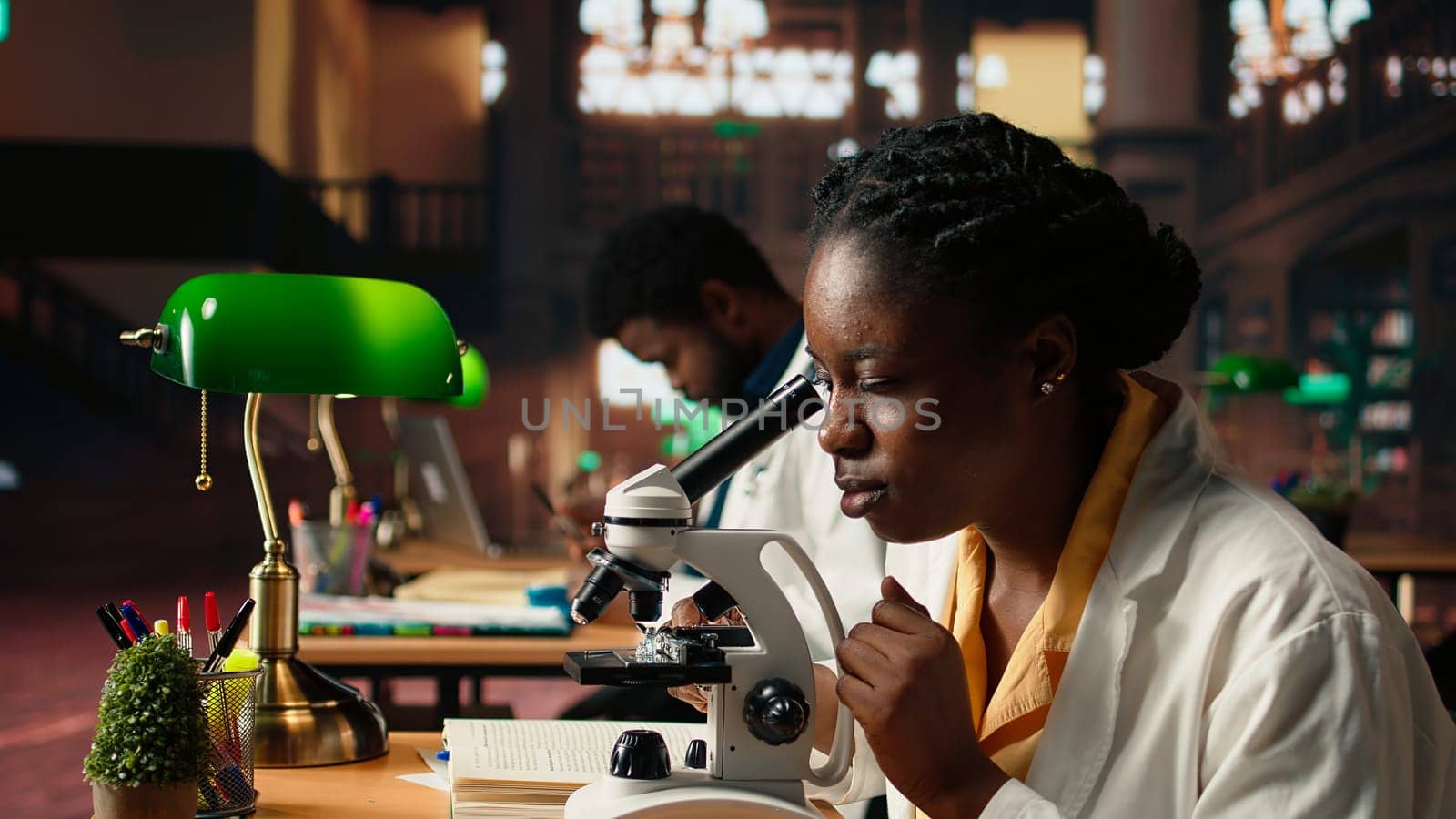 Female science student examines cells under the microscope for her scientific research in a campus library, takes notes about chemical processes and reactions for her thesis. Camera A.