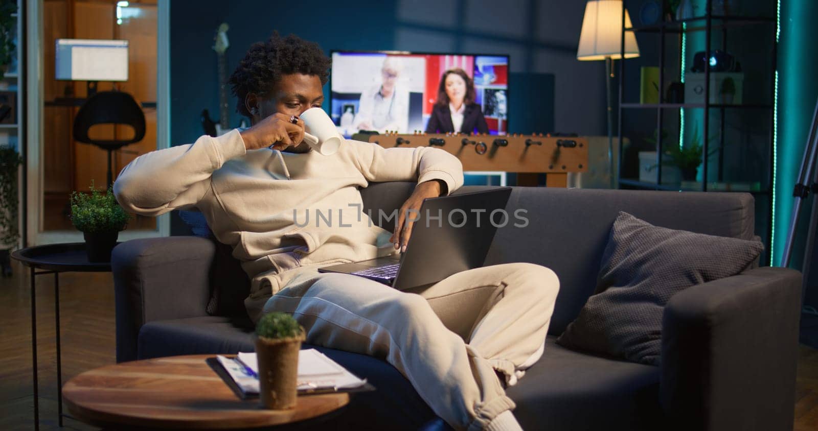 Positive african american man multitasking on a laptop in stylish loft, fully connected to the internet while managing his freelance work and remote responsibilities. Browsing web at home. Camera B.