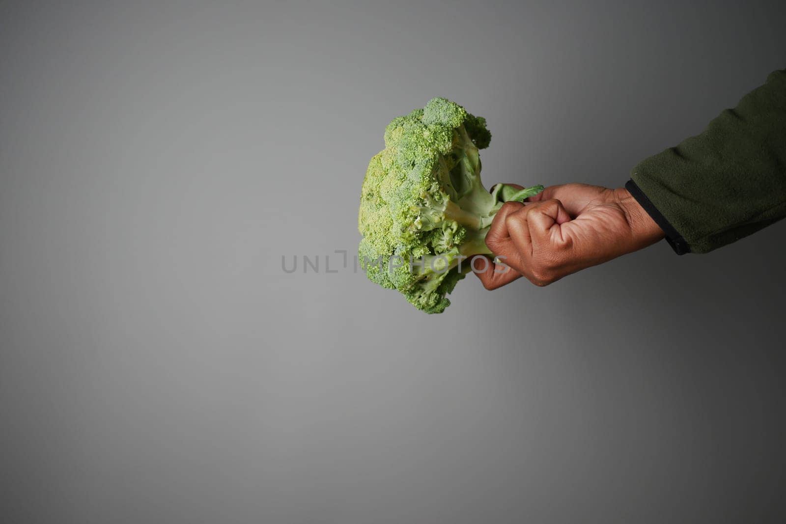 A hand grips a fresh, vibrant broccoli against a neutral gray backdrop.