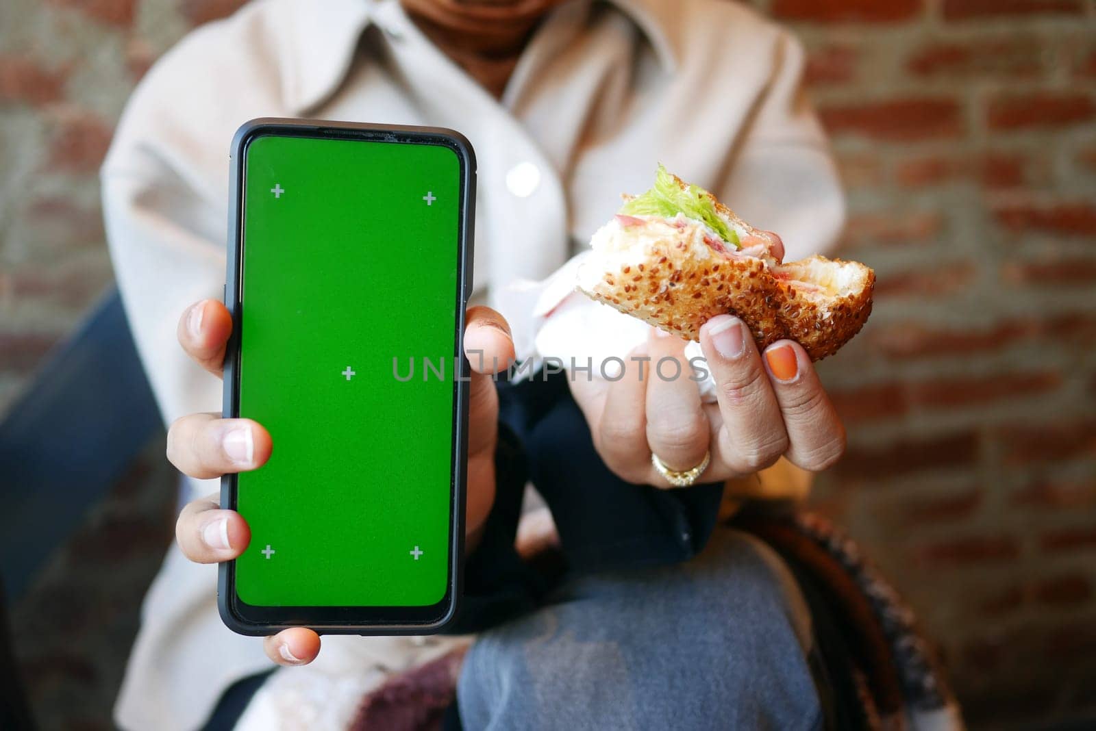 A person holds a sandwich and a smartphone with a green screen.