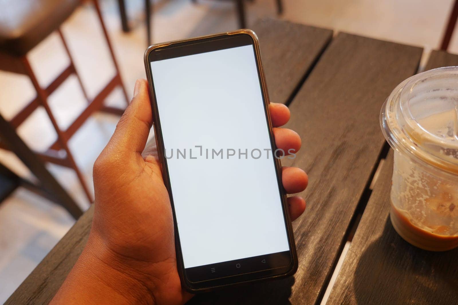 A person holds a blank smartphone next to a drink at a cafe.