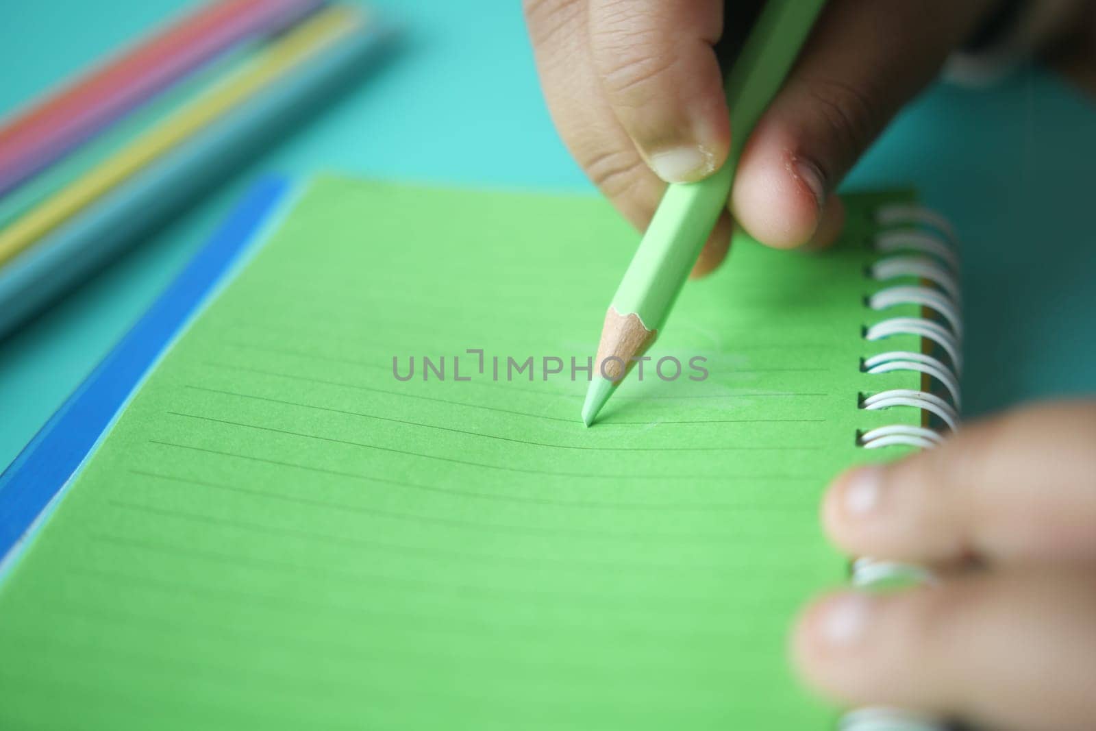 A hand holding a green pencil makes notes on a lined notebook.