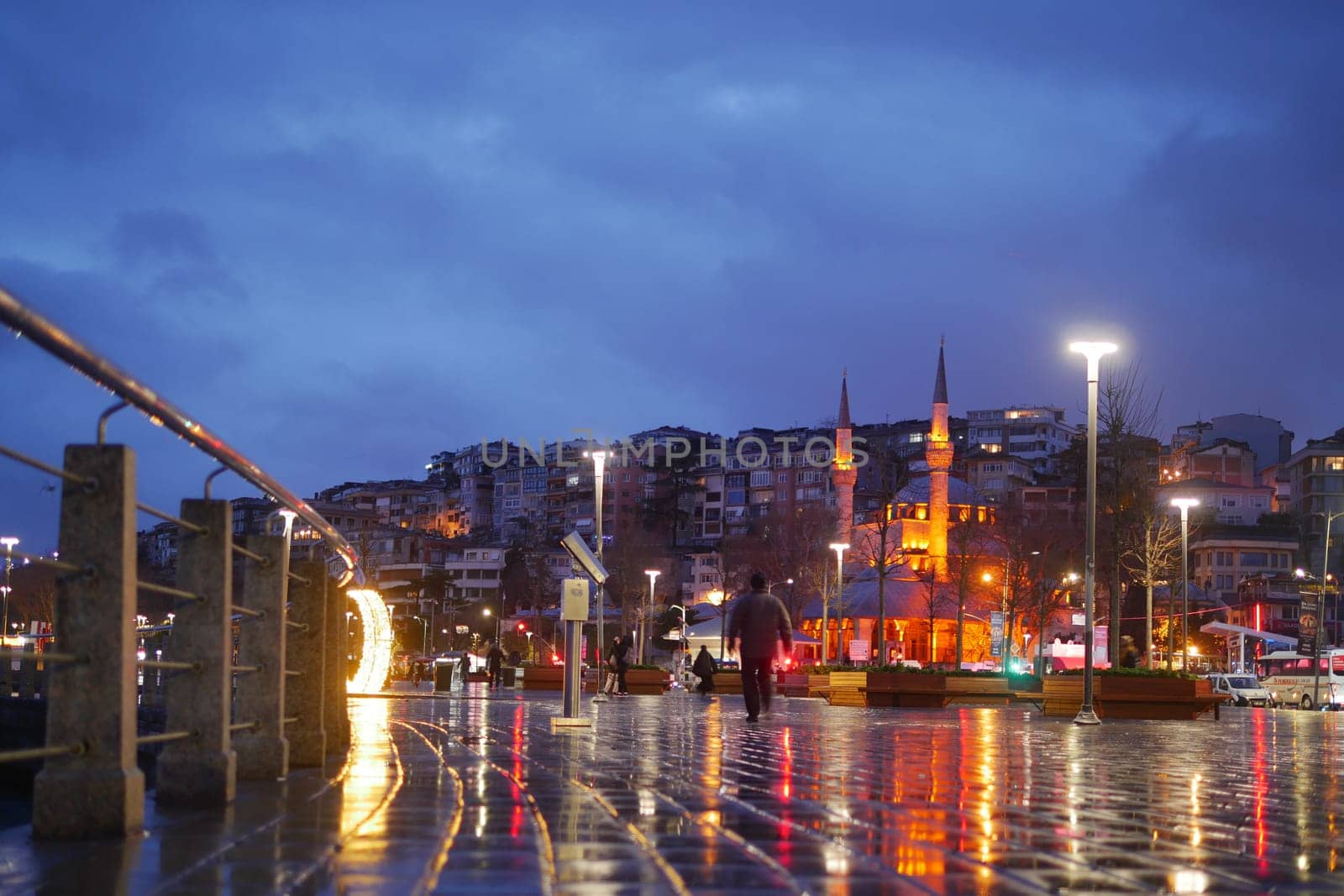 People stroll by the waterfront as lights reflect on the wet pavement.
