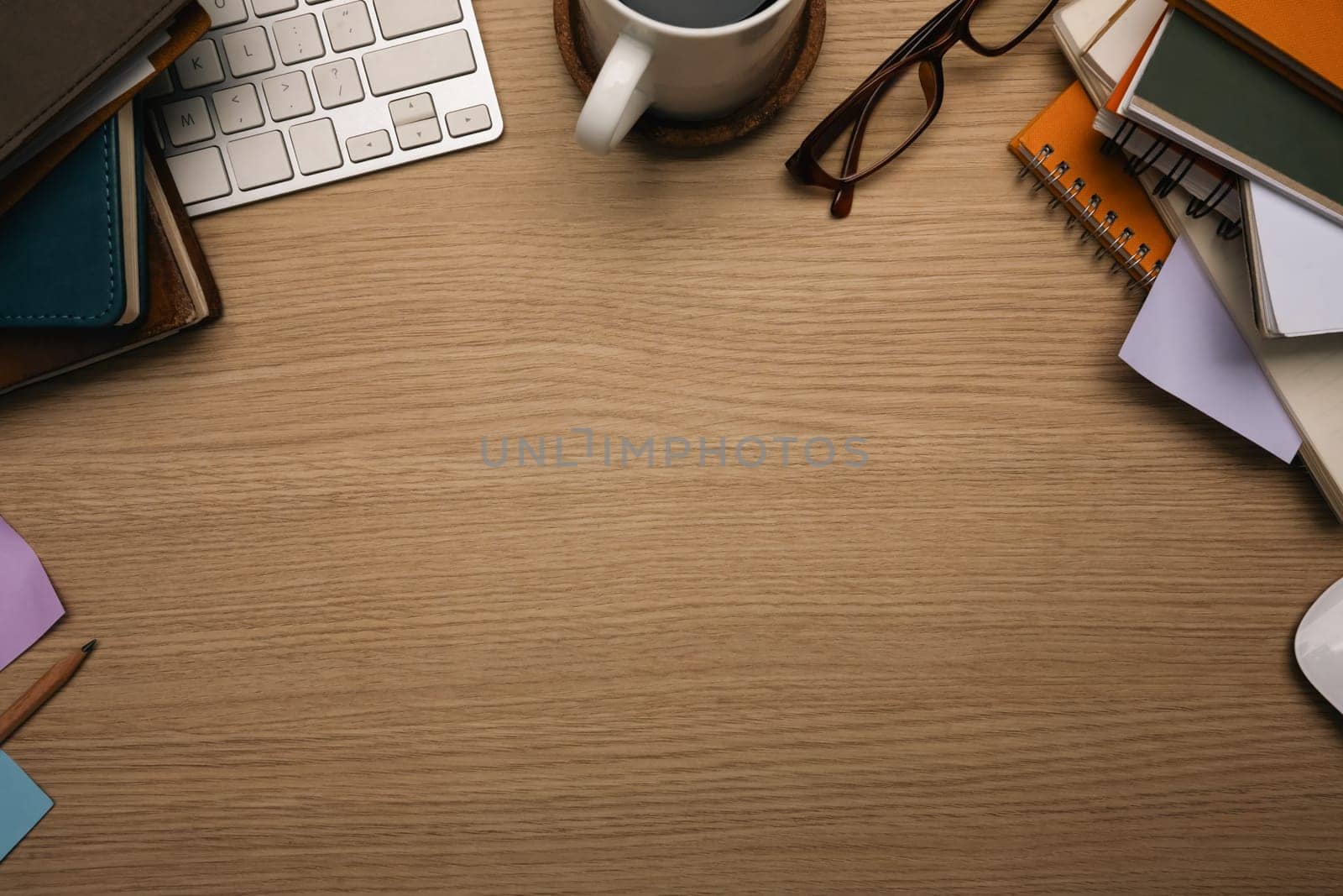 Top view of wooden desk with stacked books, a coffee cup, keyboard and glasses.