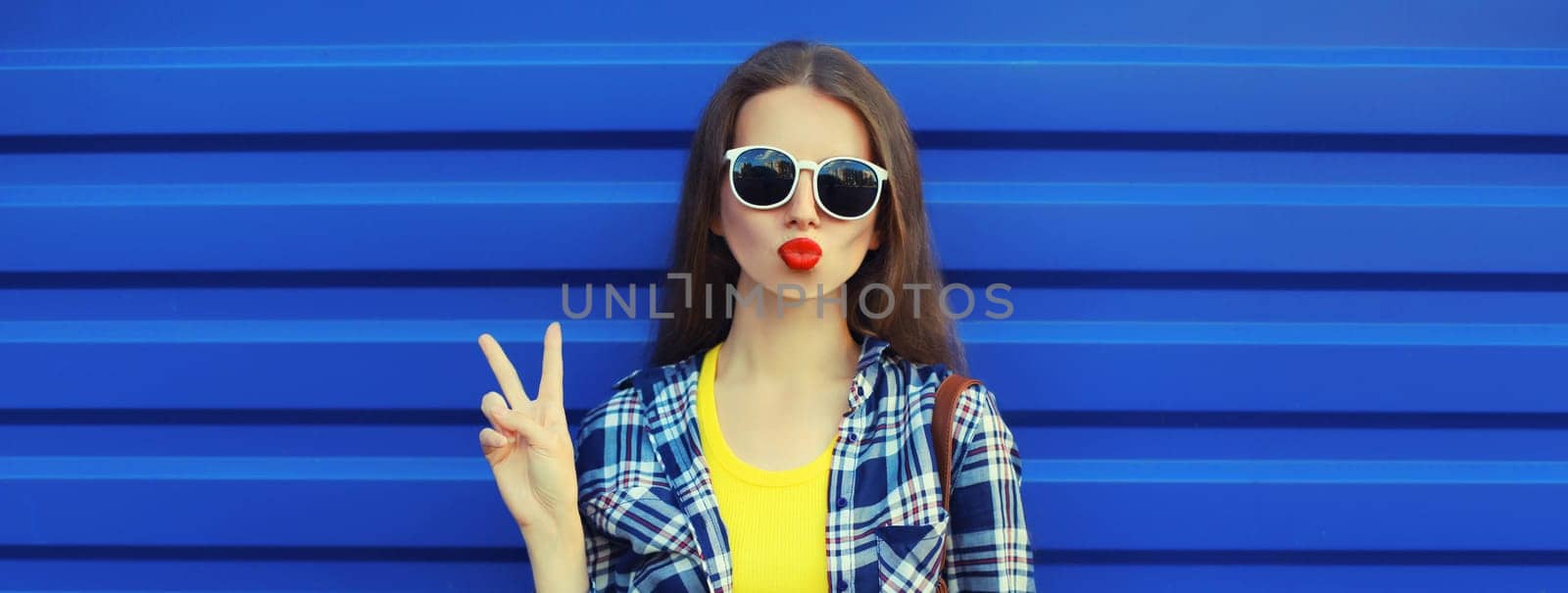 Portrait of stylish young woman blowing a kiss, cool modern girl posing in casual clothing on blue wall background