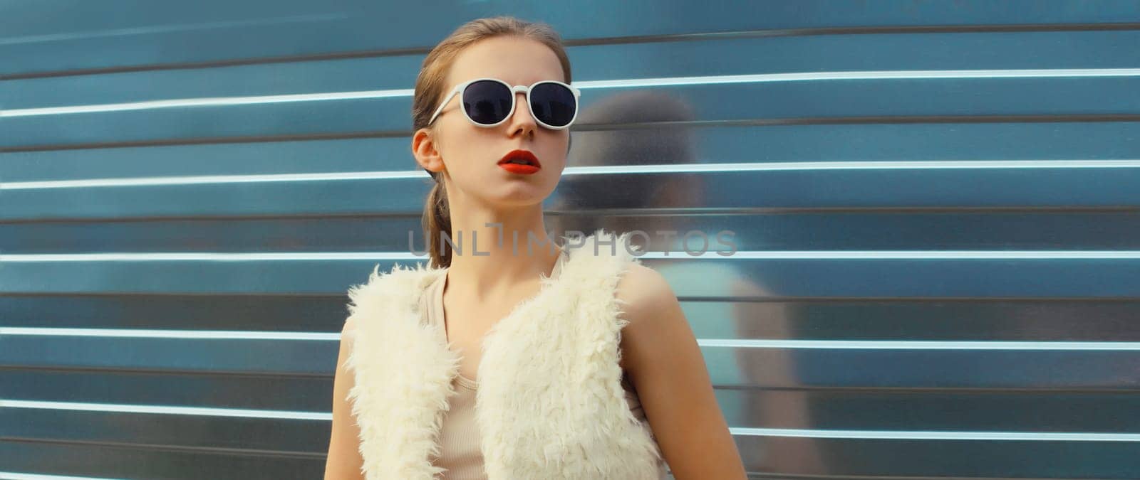 Portrait of modern stylish young woman in white fur vest, glasses, fashionable female model posing on gray silver wall background