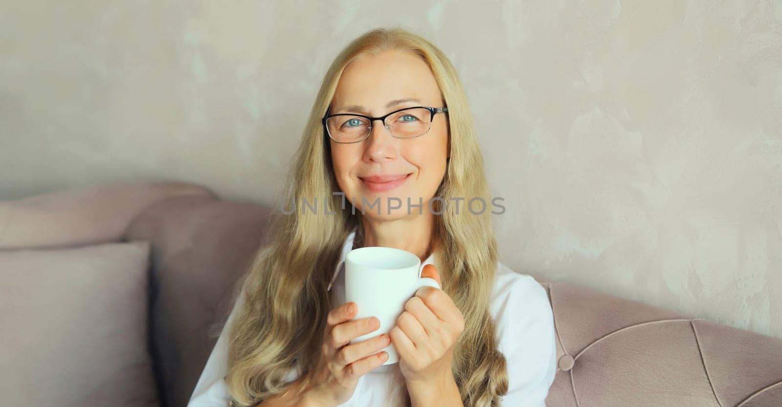 Happy satisfied mature woman enjoying hot tasty cup of coffee or tea looking at camera in early morning sitting on a couch in the living room at home