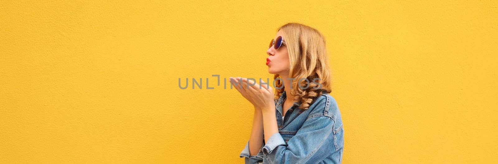 Beautiful stylish young woman blowing kiss in glasses, denim jacket posing on yellow wall background, blank copy space