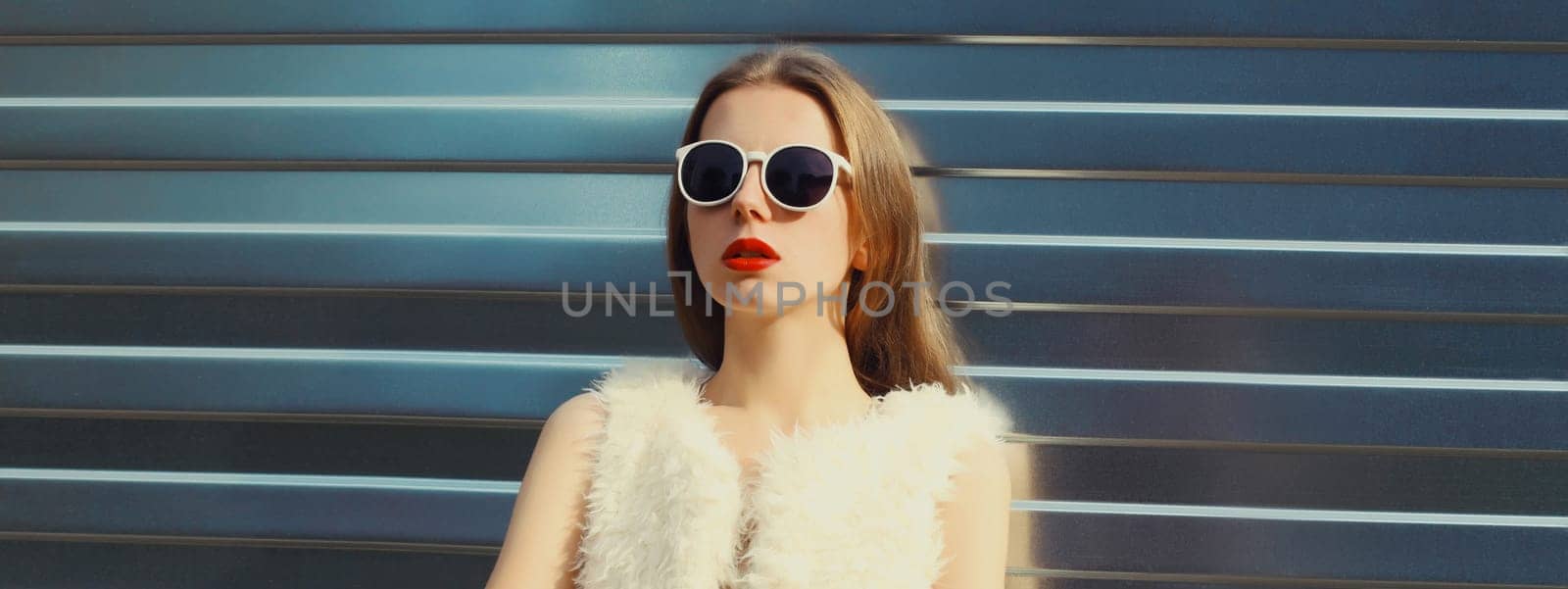Portrait of modern stylish young woman in white fur vest, glasses, fashionable female model posing on gray silver wall background