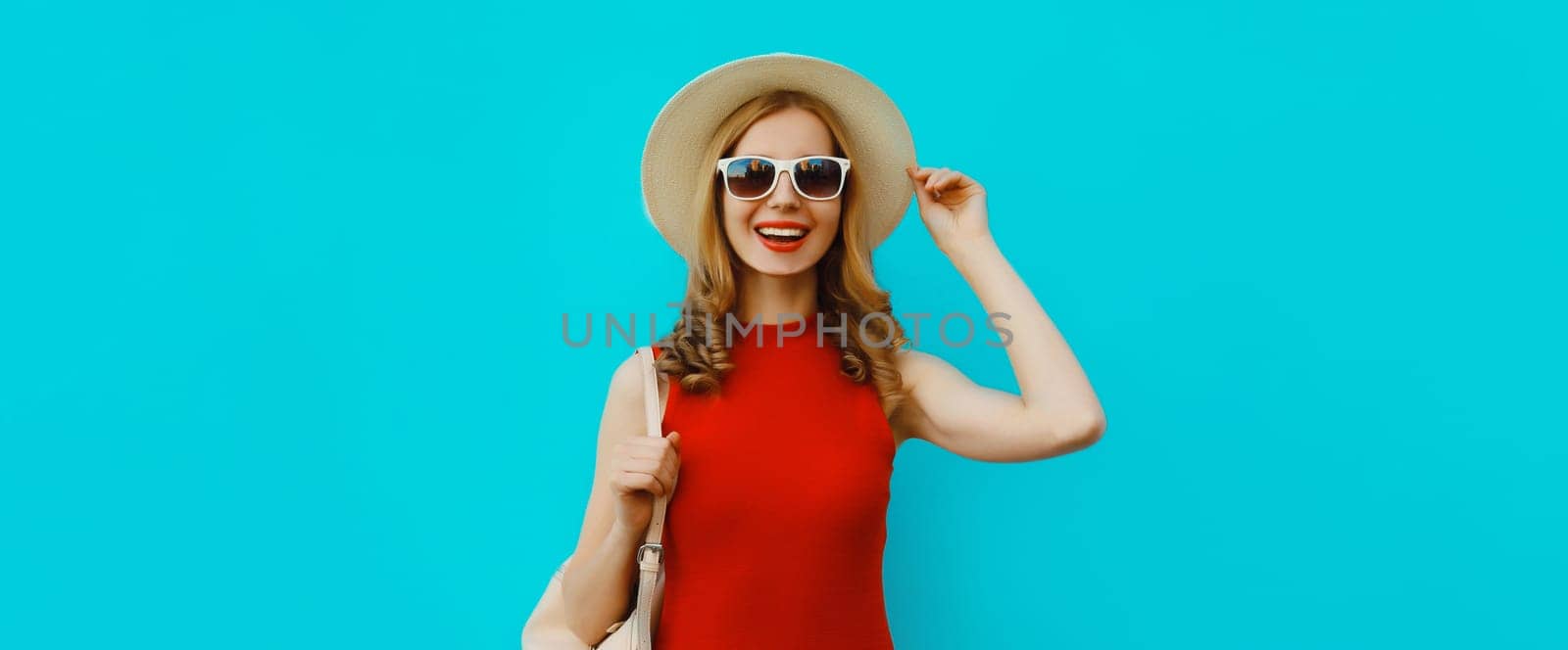 Portrait of beautiful happy young woman in summer tourist straw hat, red dress and glasses, pretty girl smiles with backpack posing on bright blue background