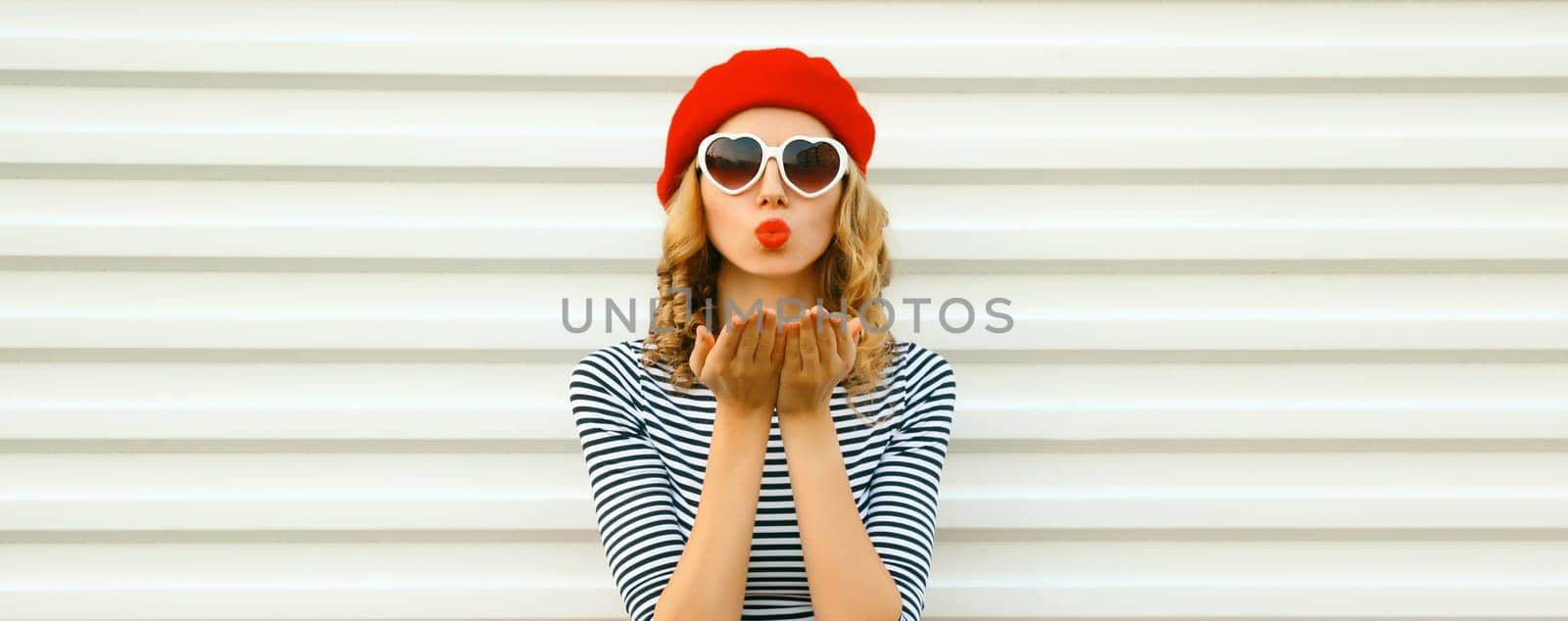 Portrait of beautiful happy young woman blowing a kiss with lipstick in red french beret, heart shaped sunglasses on white background