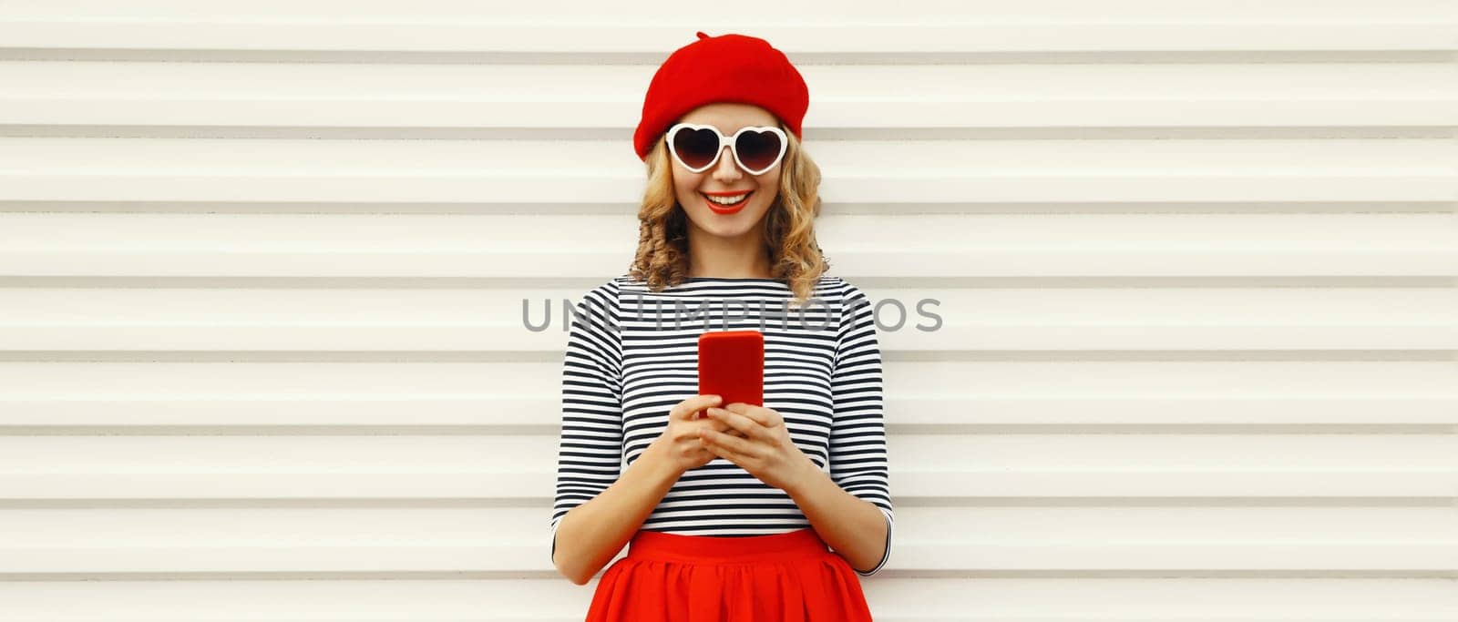 Beautiful happy smiling young woman with smartphone, stylish pretty girl holding phone in red french beret hat, heart shaped glasses on white background