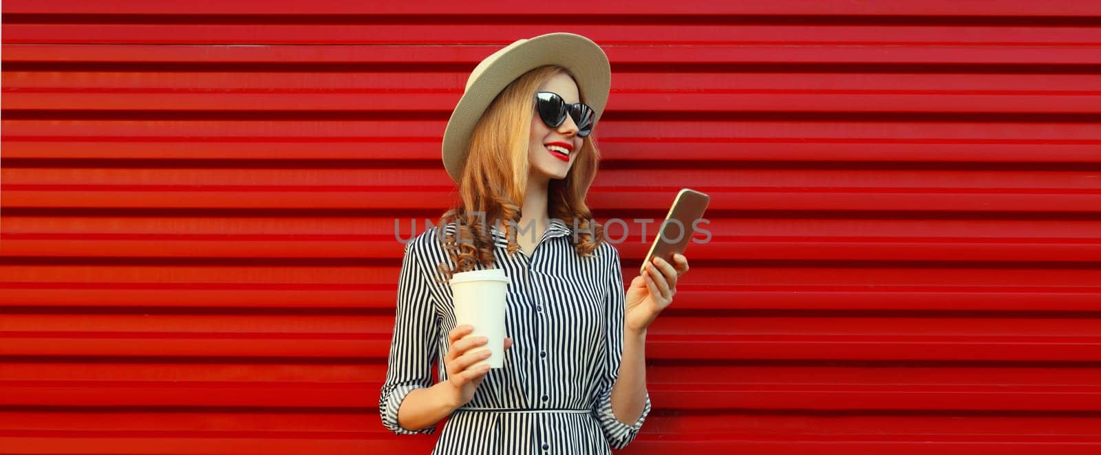 Summer portrait of stylish happy young woman with mobile phone looking at device in straw hat, holding cup of coffee on red background