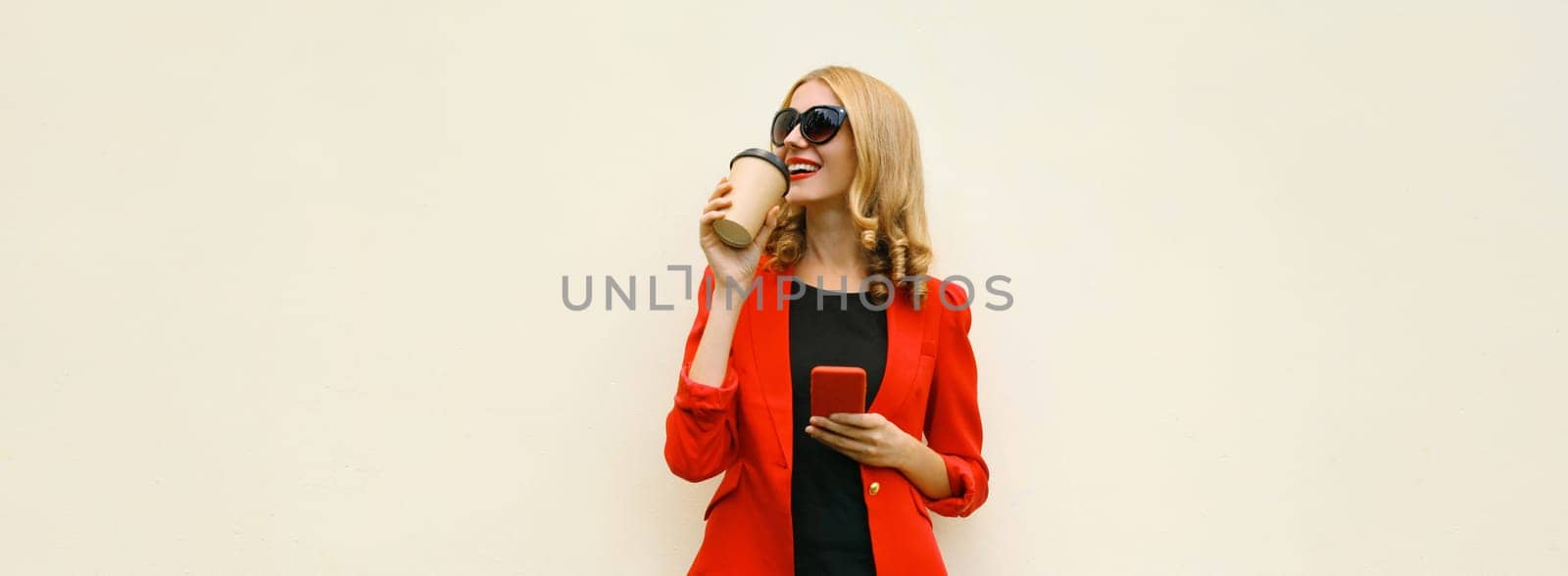 Stylish happy young businesswoman drinking coffee with phone, beautiful woman using smartphone holding cup of coffee dressed red business suit looking away standing on white background