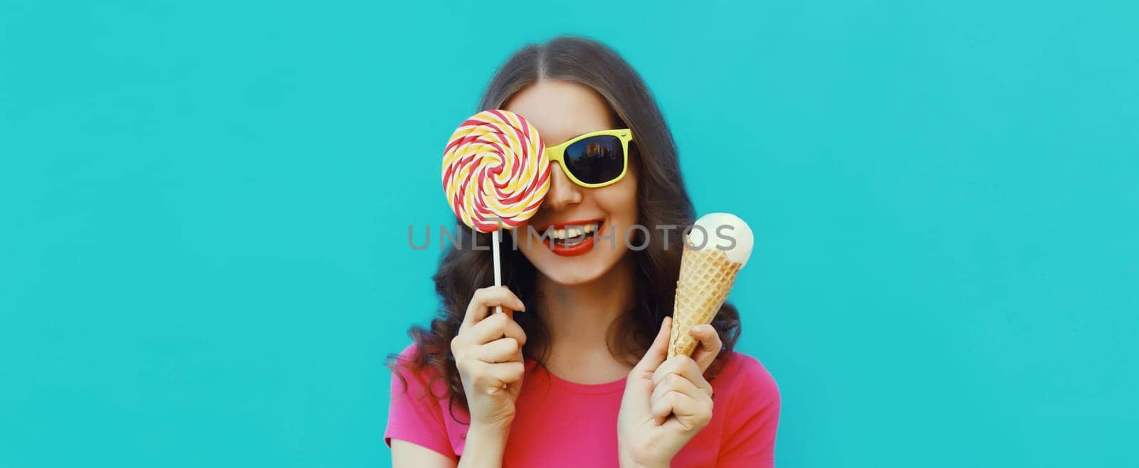 Summer portrait of happy laughing young woman holding ice cream cone and lollipop blowing a kiss on blue background