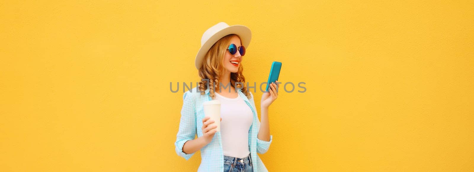 Summer stylish happy young woman with phone, joyful girl looking at smartphone holding cup of coffee dressed tourist straw hat standing on yellow background