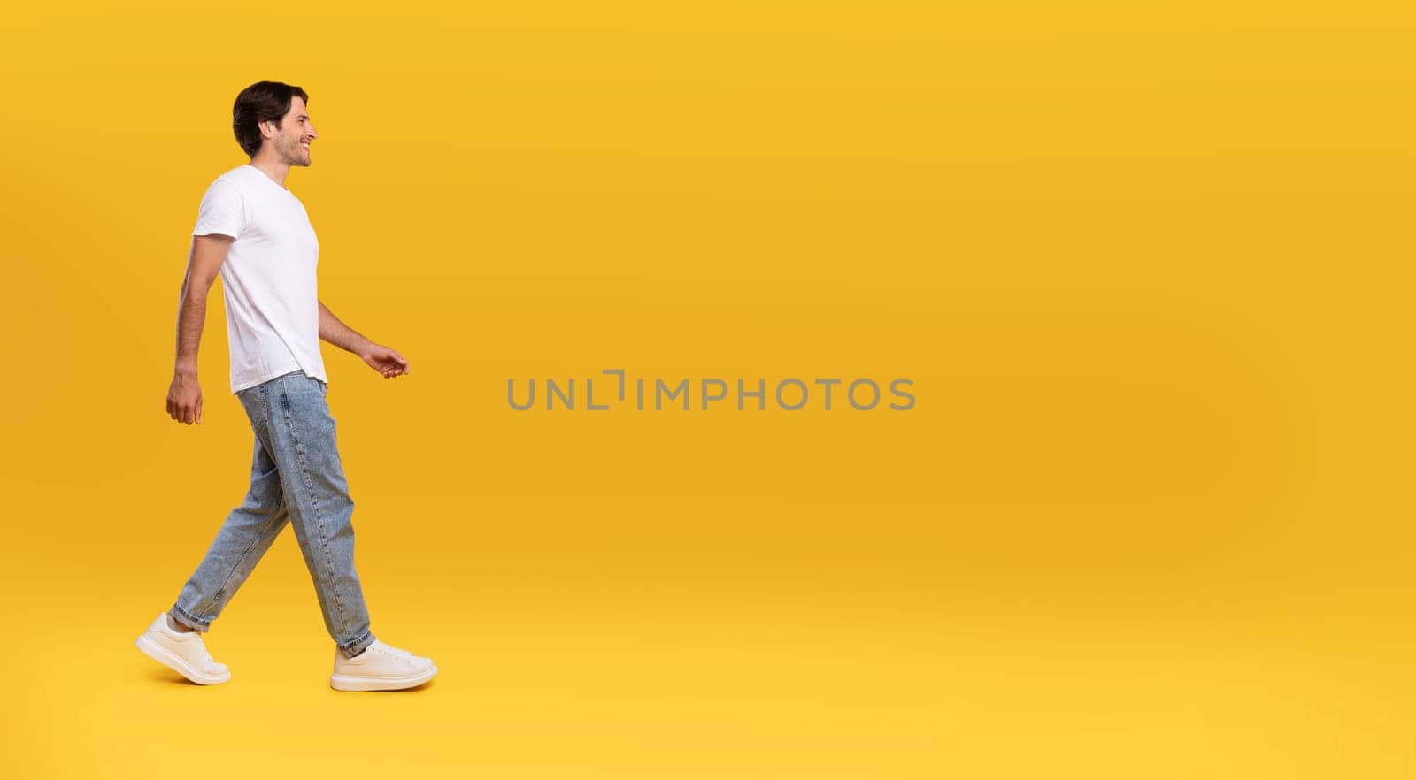 Full length body size profile side view of happy excited bearded guy in casual white t-shirt and jeans walking looking aside at free empty space isolated over orange color background, panoramic banner