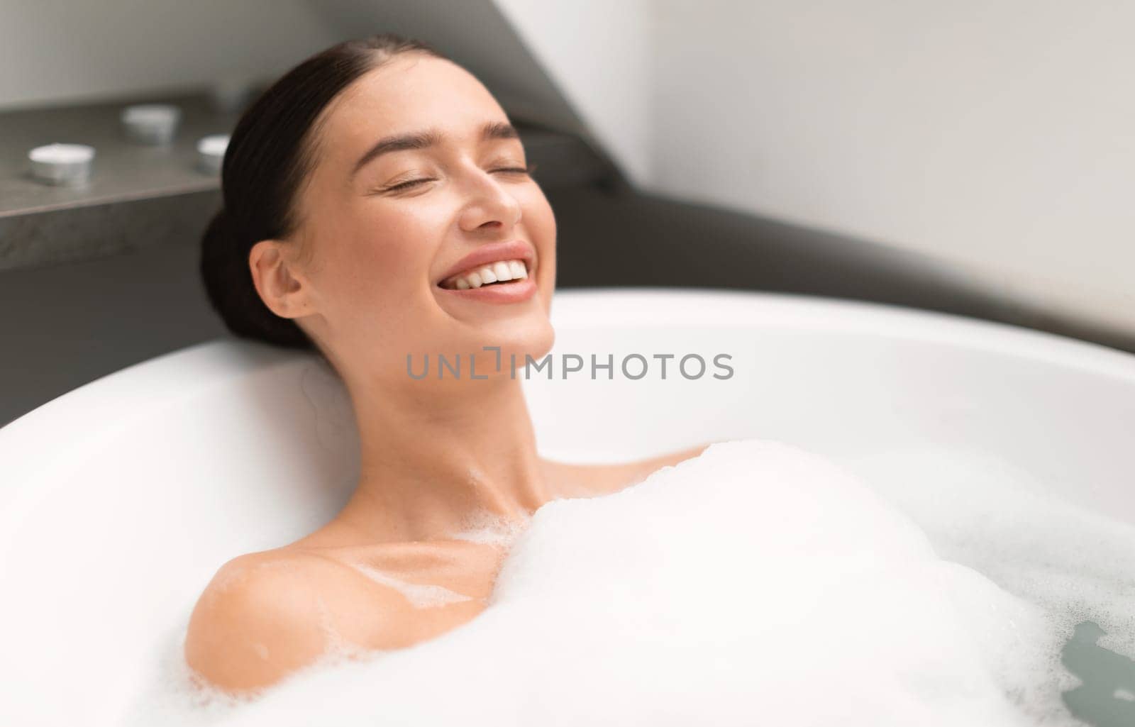 Woman Closing Eyes Taking Bath With Foam Lying In Water And Relaxing In Modern Bathroom Indoors. Gorgeous Young Female Bathing Enjoying Spa Day. Domestic Relaxation, Beauty And Wellness