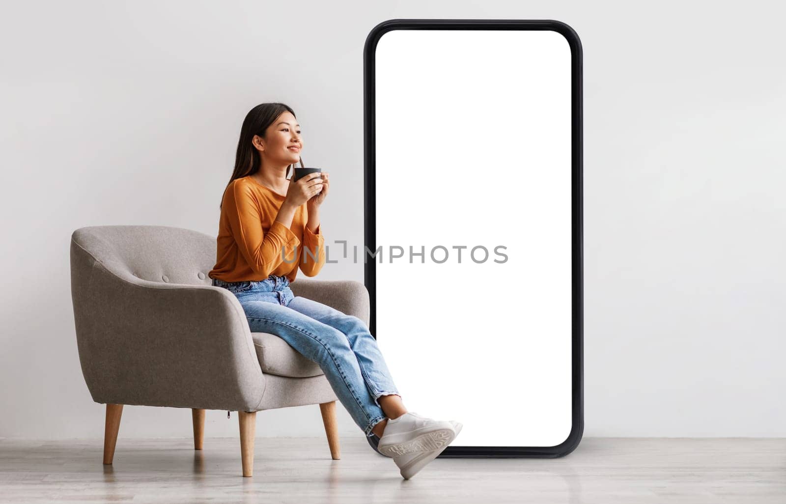 Young Asian woman drinking coffee, having break, relaxing in armchair near giant cellphone with empty screen against white studio wall, mockup for cool new mobile app or online ad design