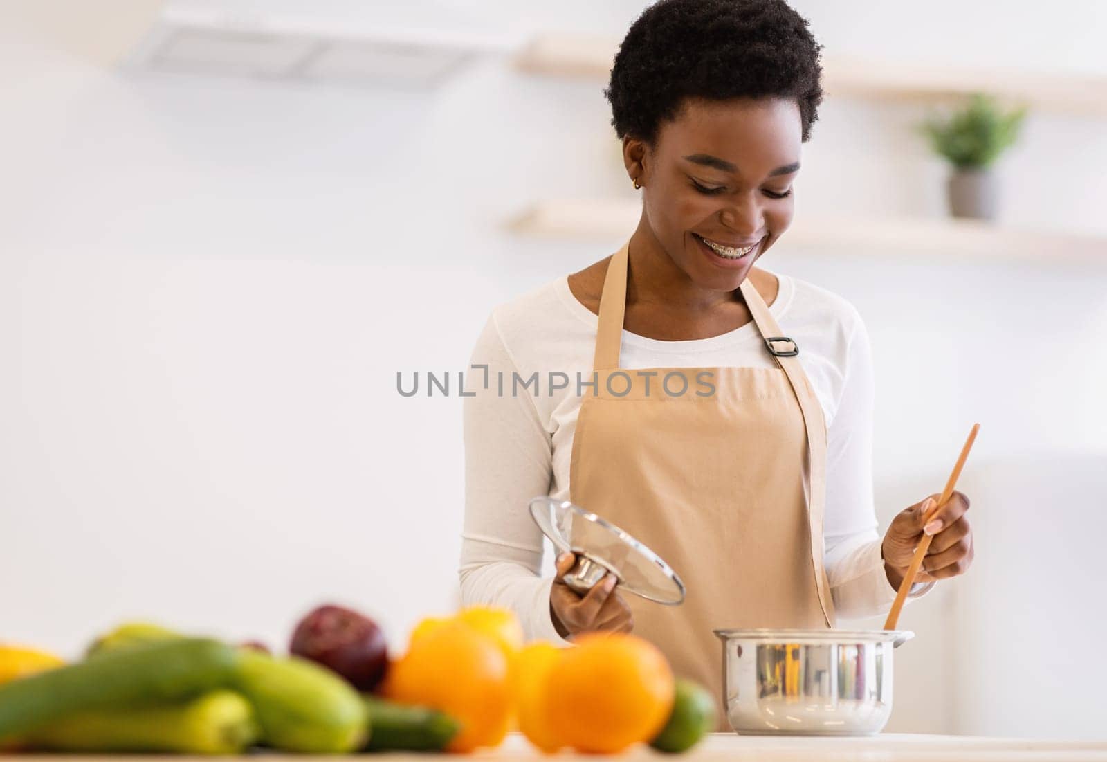 Happy African Woman Cooking Dinner In A Pot Standing In Modern Kitchen At Home, Wearing Apron. Black Housewife Preparing Meal Indoors. Healthy Food Recipes, Household And Nutrition
