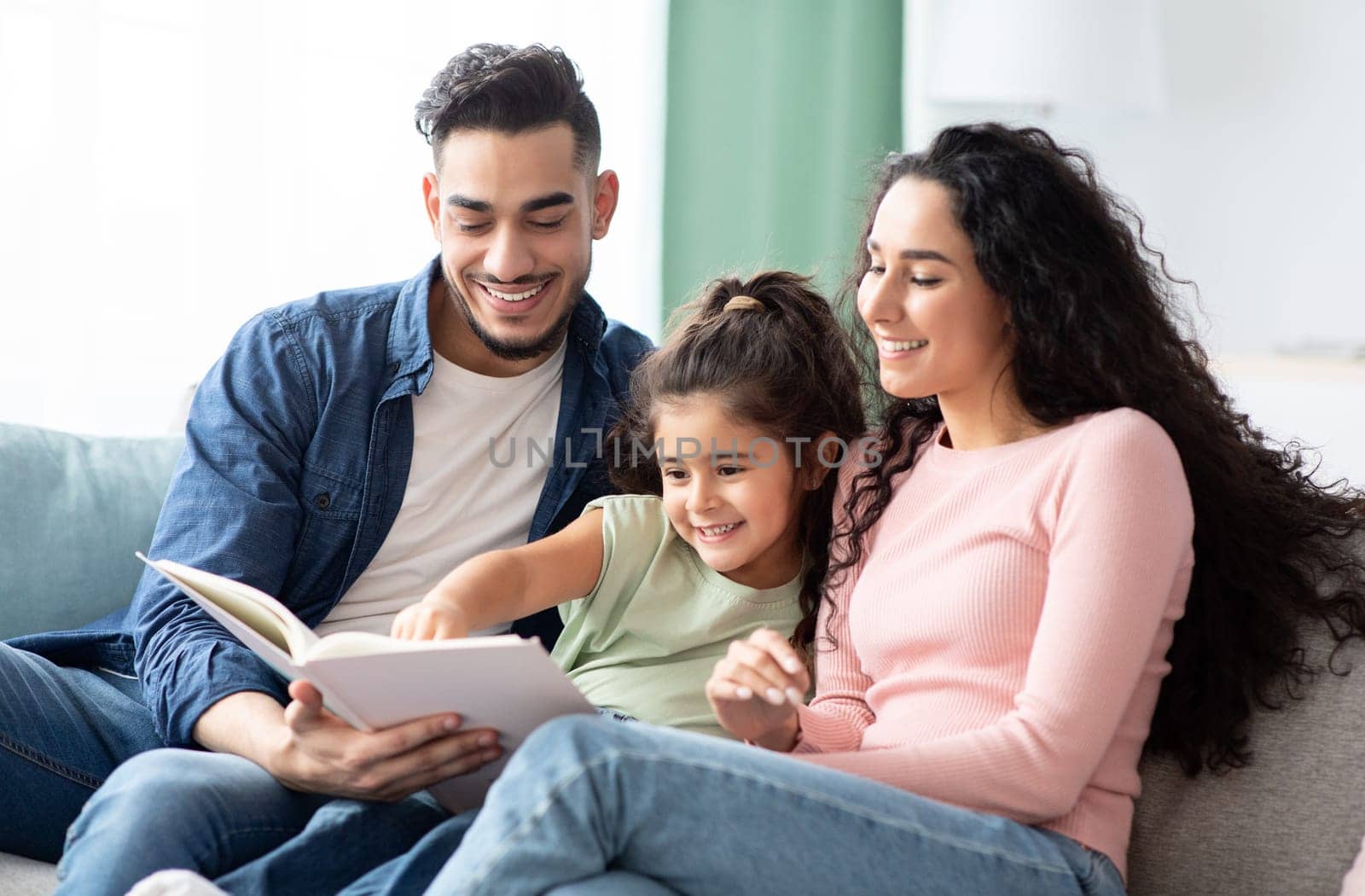 Family Time. Happy Arab Mom, Dad And Little Daughter Reading Book Together While Relaxing On Couch In Living Room, Happy Middle Eastern Parents Having Fun With Their Child At Home, Closeup Shot