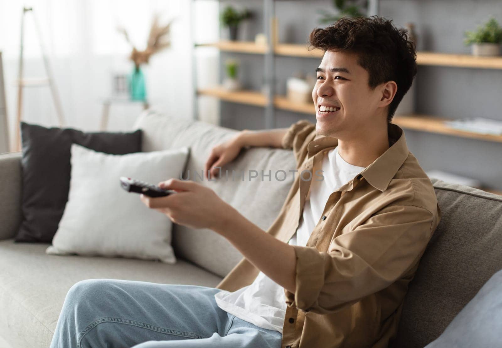 Leisure And Entertainment. Smiling asian guy watching TV show or film, holding remote control. Young man enjoying free time sitting on the comfortable couch at home in living room, free space