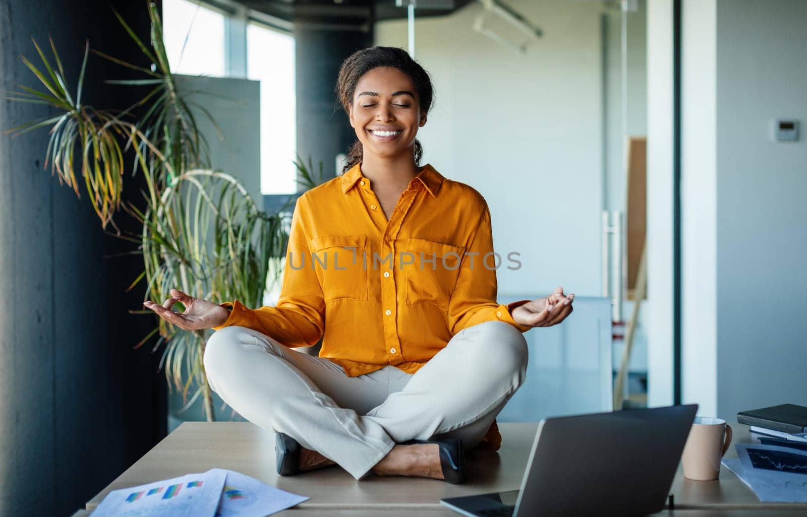 Office zen. Calm black businesswoman meditating with closed eyes on office desk, sitting at workplace in lotus position. Positive woman feeling peaceful and balanced