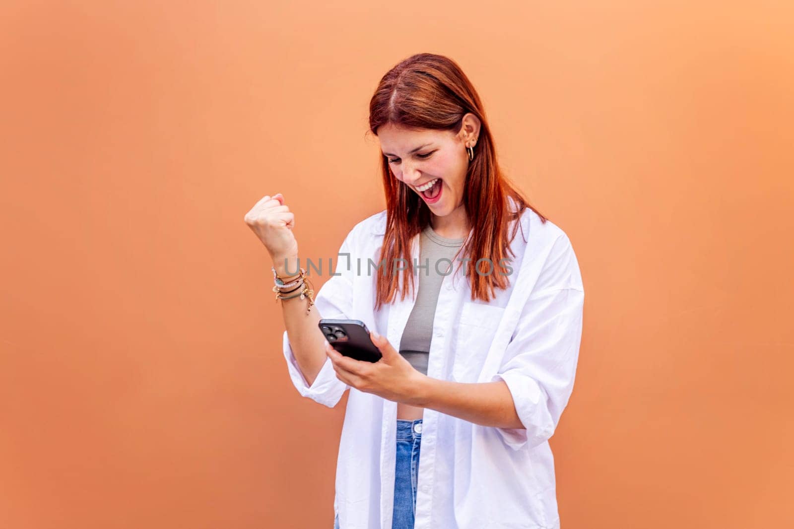young woman holding cell phone and smiling happy while celebrating with clenched fist, concept of positivity and enjoyment of technology, copyspace for text