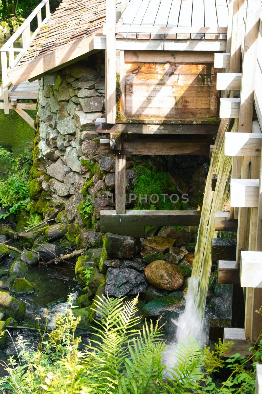 Old watermill surrounded by green nature. A small stream flows over moss-covered stones, surrounded by trees. Idyllic landscape with rustic wooden architecture.