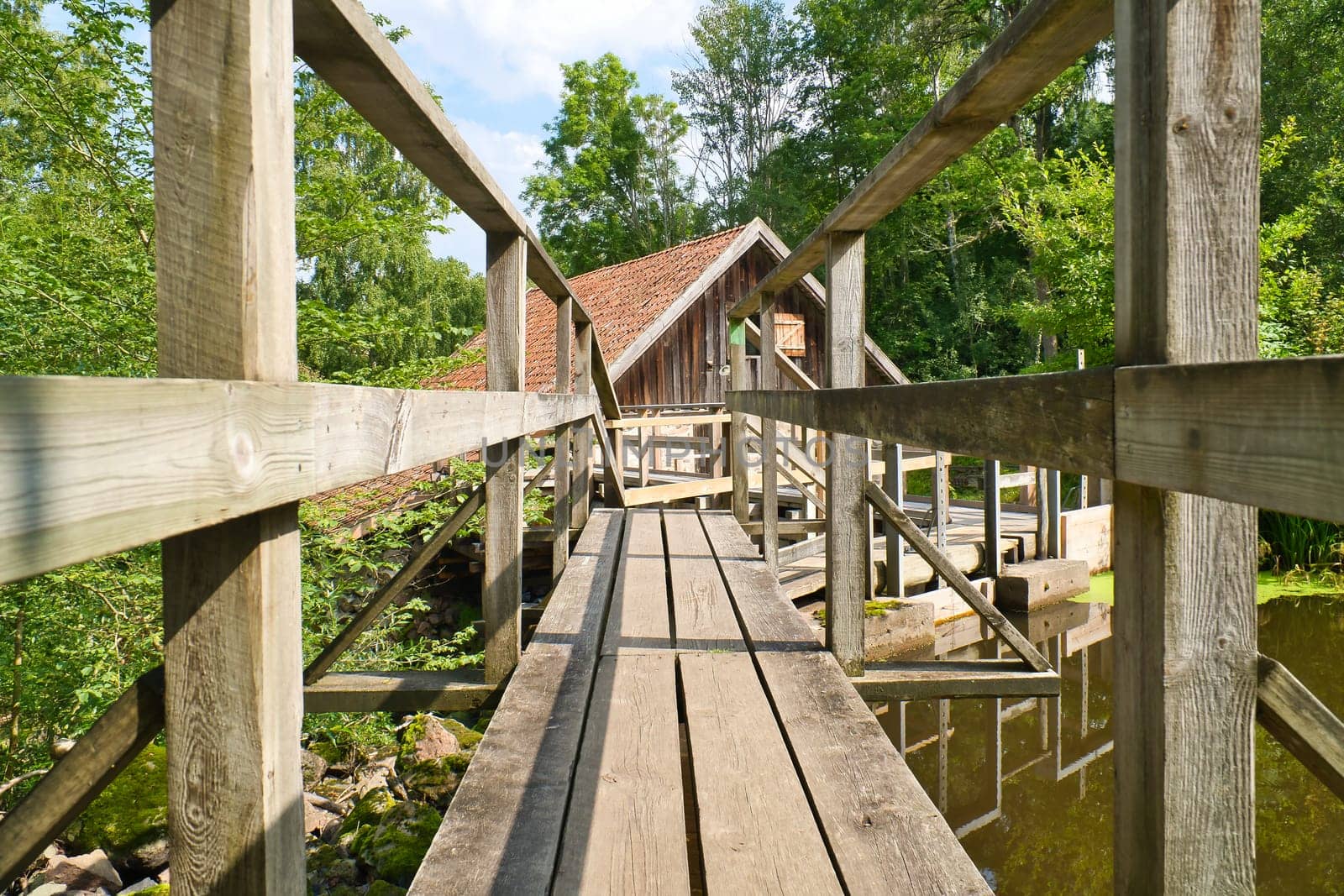 Old watermill surrounded by green nature. A small stream flows over moss-covered stones, surrounded by trees. Idyllic landscape with rustic wooden architecture.