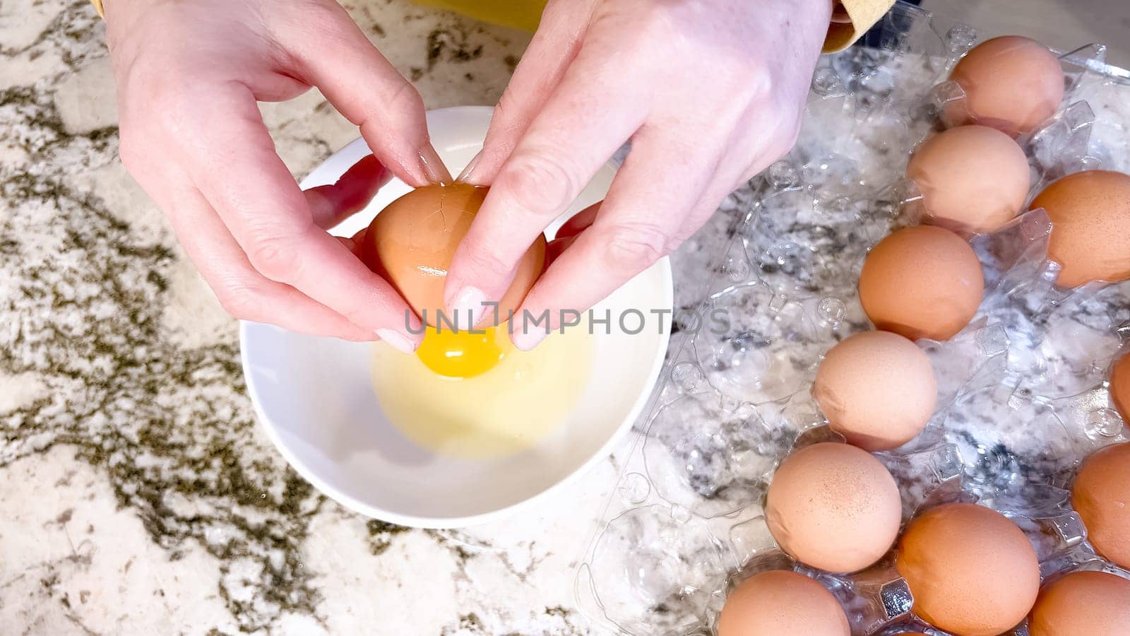 Hands are shown cracking a fresh egg open over a white plate, with a cluster of brown eggs waiting their turn on the cool, marbled surface of a well-used kitchen counter.
