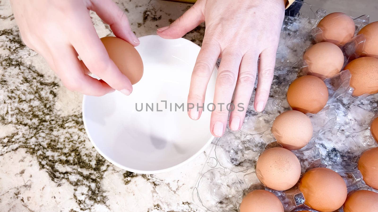 Hands are shown cracking a fresh egg open over a white plate, with a cluster of brown eggs waiting their turn on the cool, marbled surface of a well-used kitchen counter.