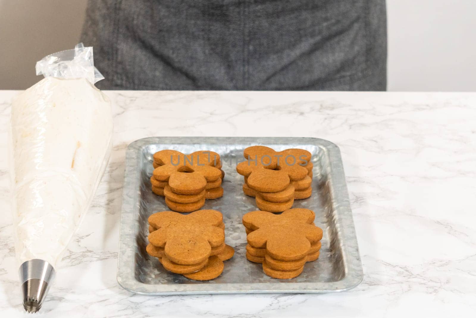 Gingerbread cookies await their second halves on a marble surface, each meticulously piped with buttercream to craft delightful sandwich treats. The precision of the piping adds a festive touch.