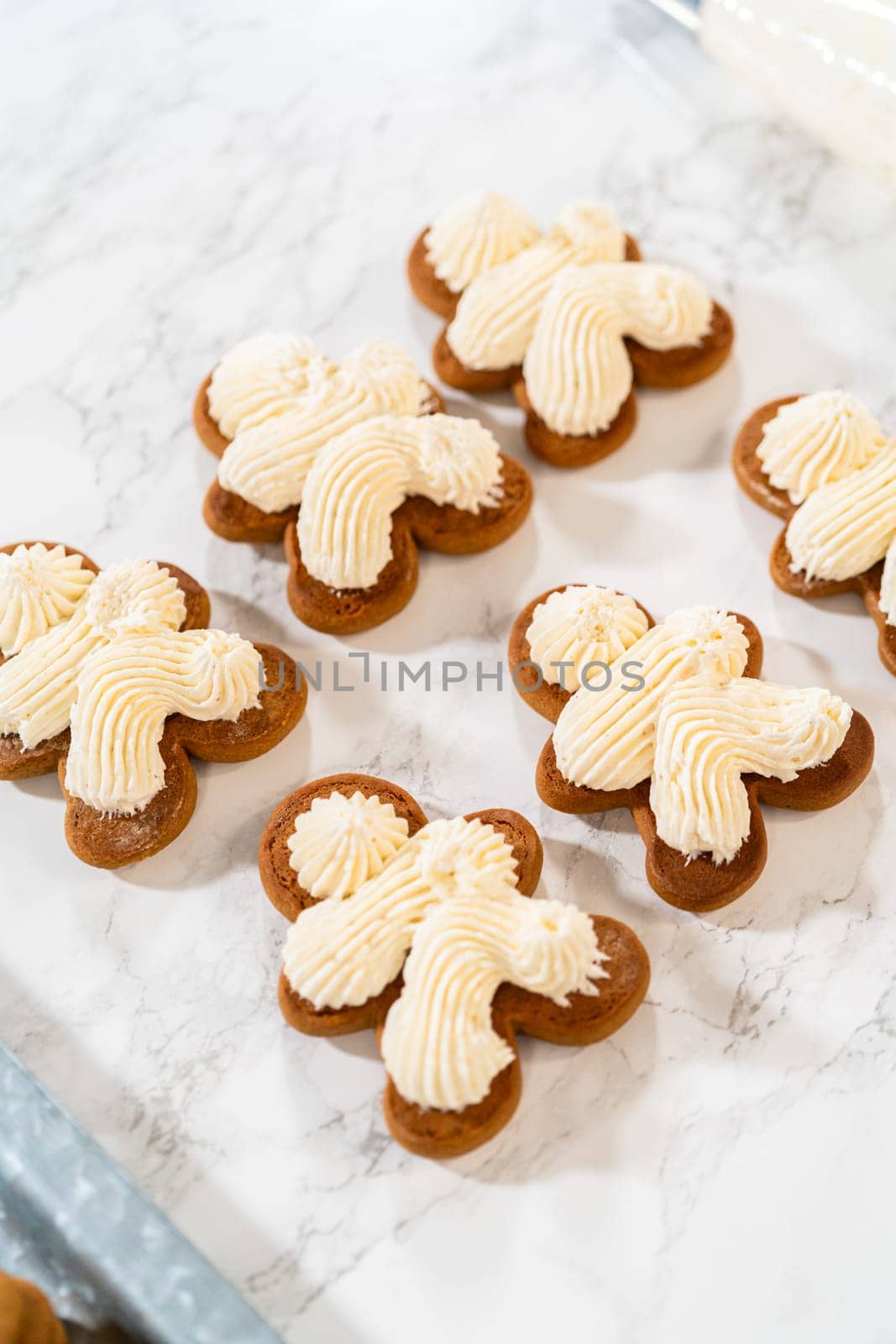 Gingerbread cookies await their second halves on a marble surface, each meticulously piped with buttercream to craft delightful sandwich treats. The precision of the piping adds a festive touch.
