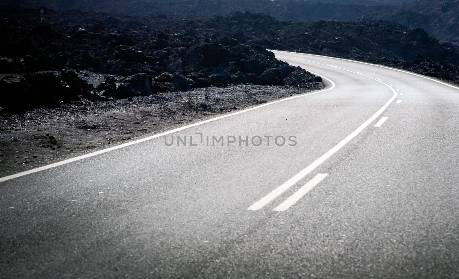 An atmospheric asphalt road among volcanic black basalt lava formations, the path is an empty scenic highway without people running through dramatic volcanic terrain with volcanoes.