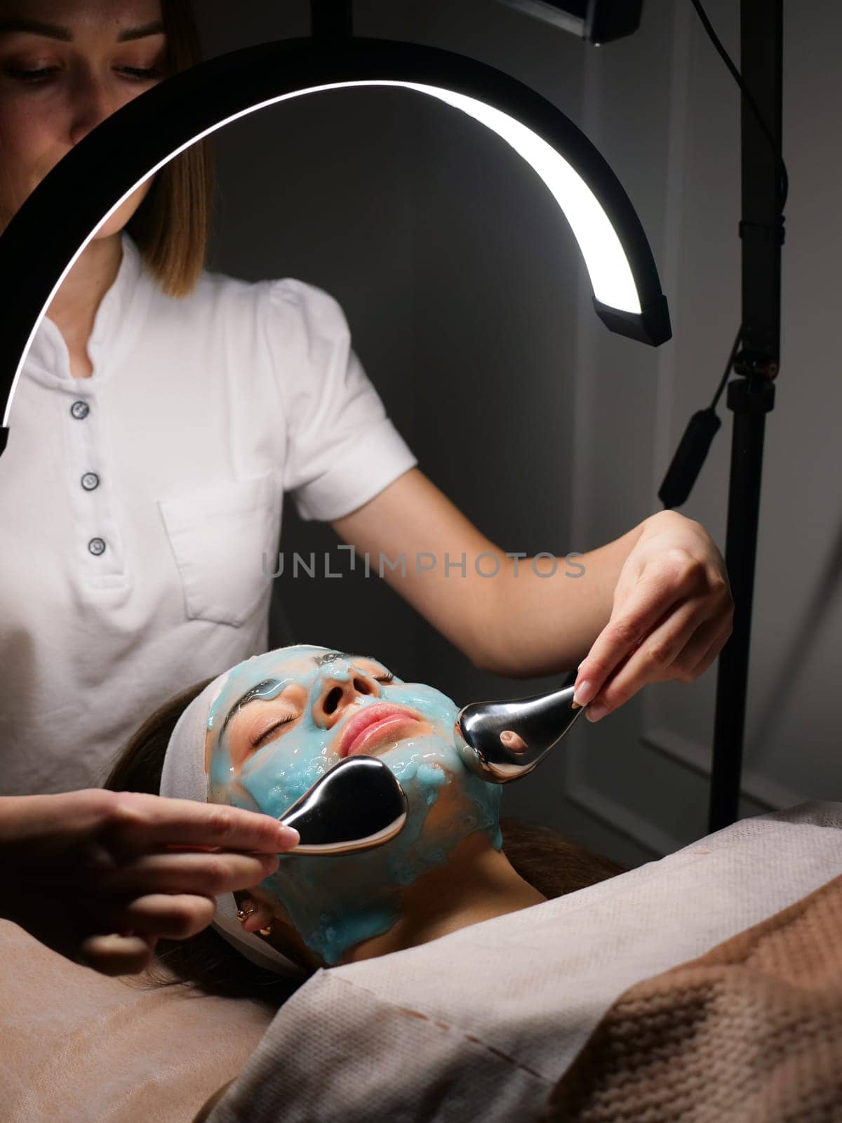 Cosmetologist does cryo massage with globules on a blue hydrogel alginate mask in the beauty salon, vertically, close-up. Woman Facial Skin and Body Care