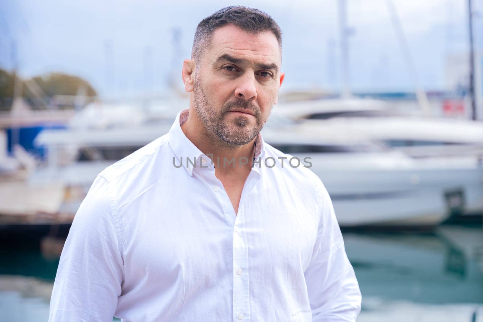 Caucasian man with short dark hair and beard stands near luxury yachts, wearing a white shirt. He looks serious and confident, gazing into the distance in a marina setting with boats and water.