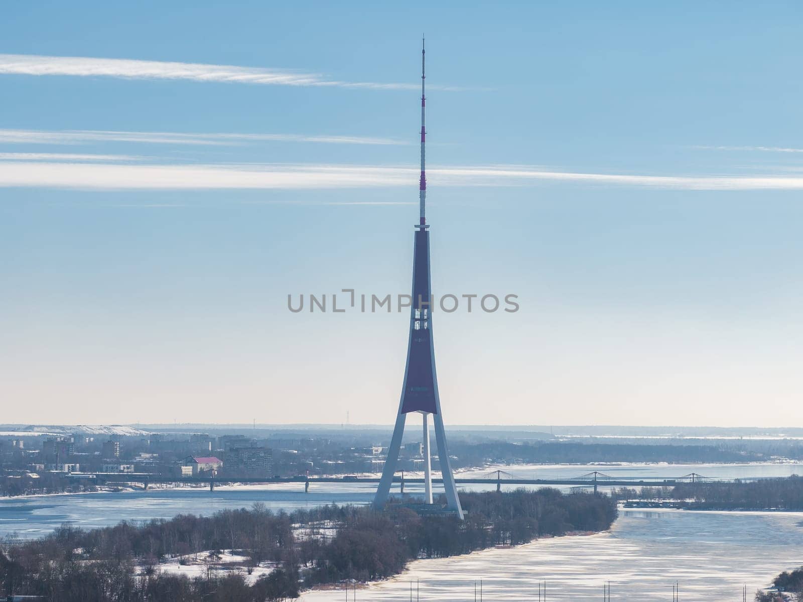 The Riga TV Tower stands on an island in the Daugava River, surrounded by snow and frozen water, with Riga's cityscape and bridges in the background.
