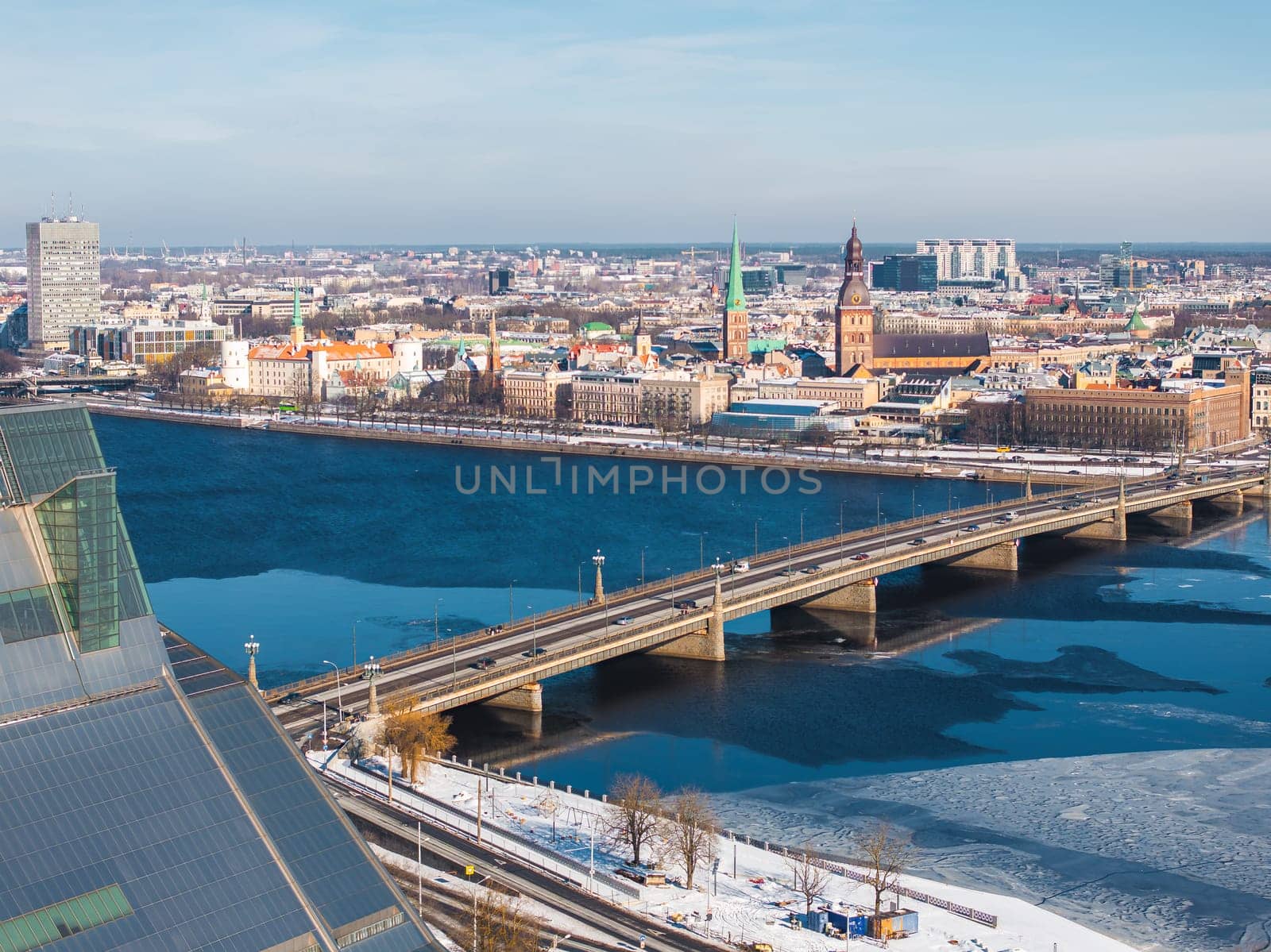 Aerial view of Riga, Latvia, featuring the Daugava River, National Library, Old Town landmarks, a bridge, and floating ice under a clear blue sky.