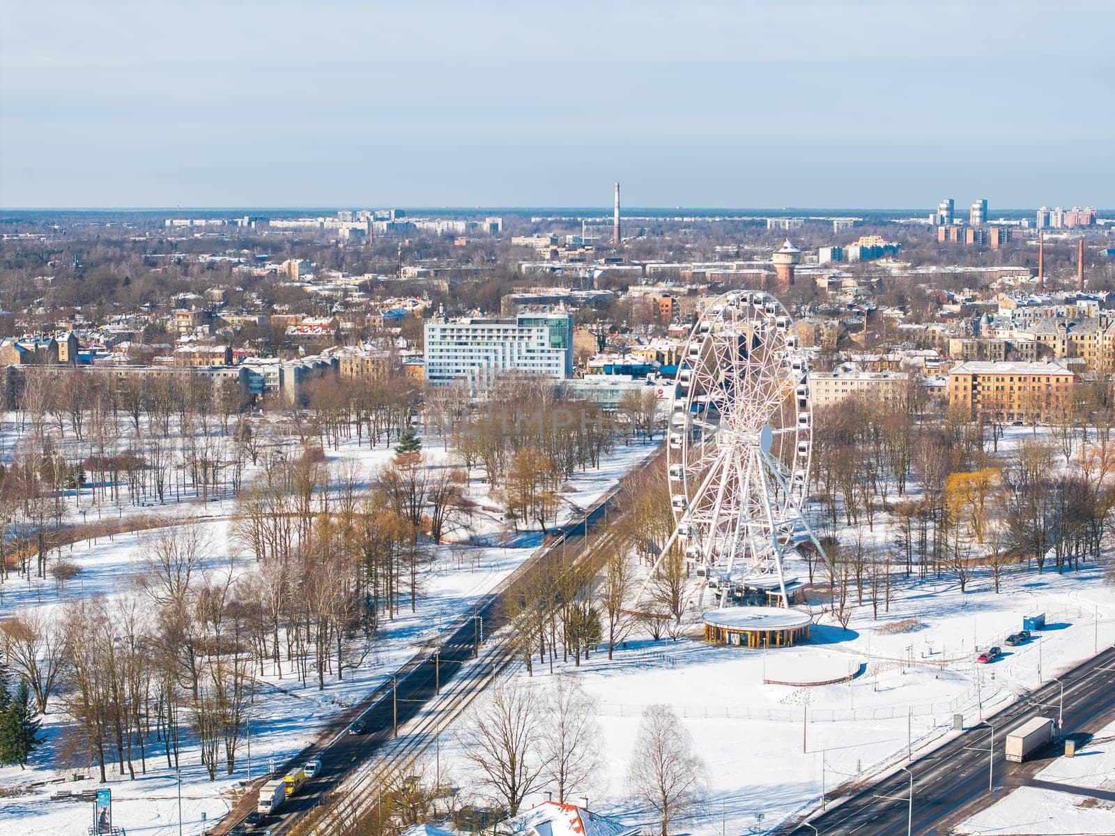 Aerial view of Riga, Latvia, featuring a Ferris wheel in a snowy park, urban buildings, tram tracks, and a clear sky with distant cityscape elements.