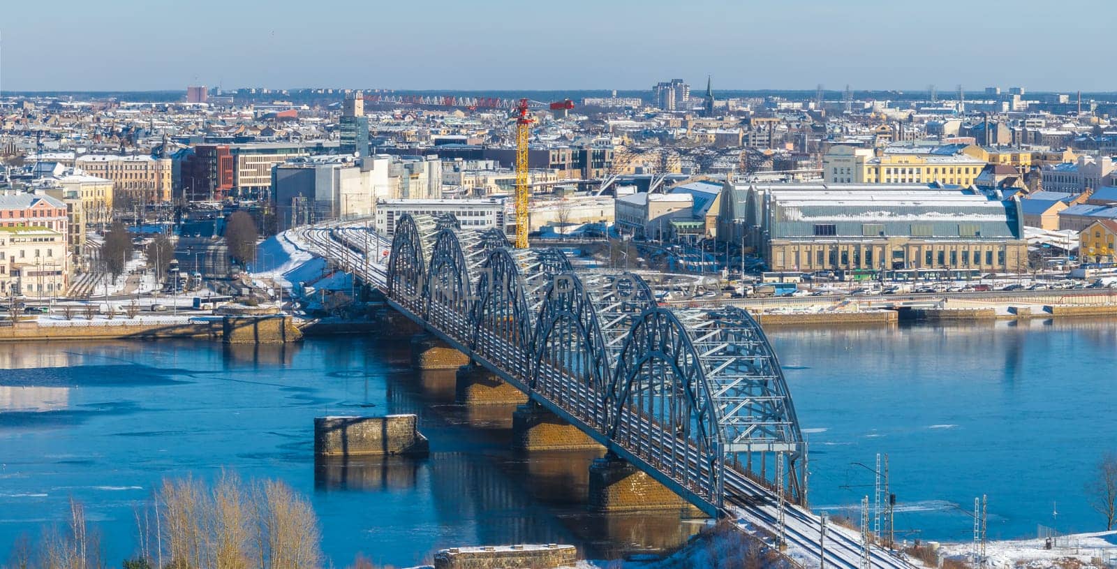 Aerial view of Riga, Latvia, in winter featuring the Railway Bridge over the Daugava River, Central Market pavilions, and a mix of historic and modern buildings.